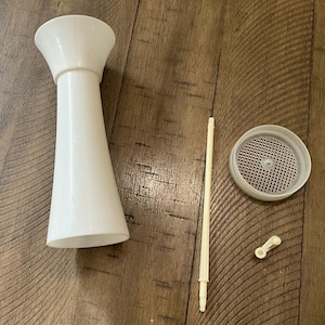 May include: A disassembled white plastic kitchen tool, possibly a food mill. The parts include a tall, conical body, a perforated lid, a long rod, and a small handle. The components are arranged on a wooden surface.