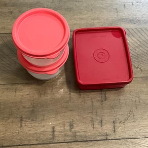 Op de afbeelding: Een stapel van drie kleine, transparante containers met koraalkleurige deksels, naast een enkele vierkante container met een bijpassend deksel. De deksels hebben een cirkelvormig ontwerp. De containers staan op een houten oppervlak.