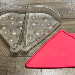 May include: Clear plastic pizza slice container with a red lid. The container is made of two clear, triangular pieces that snap together. The red lid is also triangular and fits over the top of the container. The container has a series of small, round bumps on the inside.