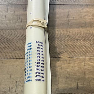 Peut inclure: Un tapis de mesure Tupperware blanc enroulé avec une attache beige. Le tapis affiche des mesures en cuillères à café, onces liquides, pintes et millilitres. Le logo Tupperware est visible en haut.