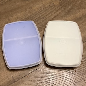 May include: Two plastic food storage containers, one is light blue and the other is white. Both containers have a divided section in the middle.