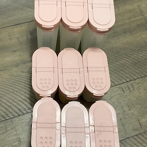 Op de afbeelding: Een verzameling van negen roze kruidenpotten met doorzichtige plastic lichamen. Elke pot heeft een klapdeksel met een schud- en schenkoptie. De potten zijn gerangschikt op een houten oppervlak.