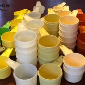 May include: A collection of plastic measuring cups in various colours including white, yellow, green, orange, and tan. The cups are arranged in stacks and individual units. Text in the upper left corner reads: Replacement pieces, Choose colour/size.