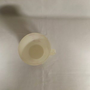 以下が含まれることがあります： 小さなオフホワイトの計量カップを真上から見た写真。取っ手が付いています。カップは円形の底とわずかに広がった縁があります。側面に測定マークが見えます。カップは半透明の素材でできています。