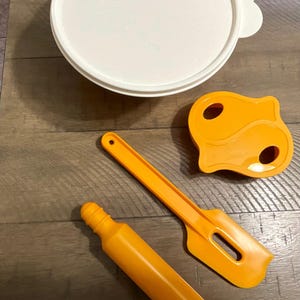 Könnte beinhalten: Eine Sammlung von orangefarbenen und weißen Küchenutensilien, darunter ein runder Deckel mit Griff, ein Nudelholz, ein Spatel und ein einzigartig geformtes Werkzeug. Die Artikel sind auf einer Holzoberfläche angeordnet, was auf ein Back- oder Essenszubereitungsthema hindeutet.