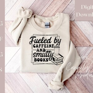 Puede incluir: Una sudadera beige con el texto "Fueled by Caffeine and Smutty Books" en letras negras. El texto est&aacute; rodeado por un marco cuadrado negro. Una ilustraci&oacute;n en blanco y negro de una taza de caf&eacute; y una pila de libros est&aacute; dentro del marco.