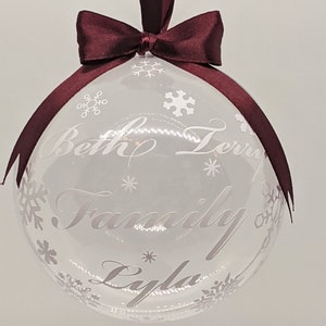 May include: Clear glass Christmas bauble with a burgundy ribbon and bow. The bauble is decorated with white snowflakes and the text "Beth Terry Family Lyla".