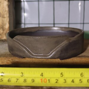 May include: A small, dark gray ceramic ashtray with a unique design. The ashtray is approximately 5 inches in diameter. A yellow tape measure is in the foreground.