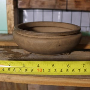 May include: A round, brown ceramic bowl with a wide rim. A yellow tape measure is placed next to the bowl, showing measurements in inches. The bowl appears to be approximately 7 inches in diameter.
