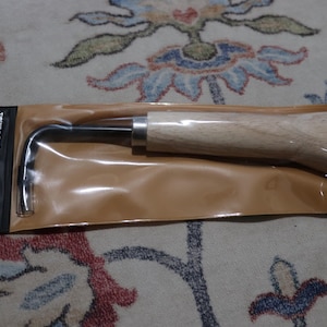 May include: A bonsai tool with a wooden handle and a curved metal end, packaged in a clear plastic sleeve. The tool has a black metal shaft and a silver metal collar where the handle meets the shaft. The packaging includes a black label with white text.