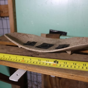 May include: A beige, oblong decorative bowl with three black square accents. A yellow tape measure shows a length of 16 inches. The bowl is on a wooden shelf, with a green wall in the background.