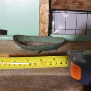 May include: A small, green ceramic dish with three small feet. The dish is approximately 5 inches long, as measured by a yellow tape measure. The dish is on a wooden surface.