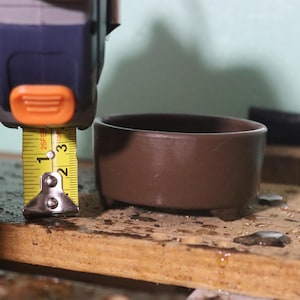 May include: A brown ceramic bowl, approximately 2 inches in diameter, is measured with a yellow tape measure. The bowl is sitting on a wooden surface. The tape measure is held by a person.