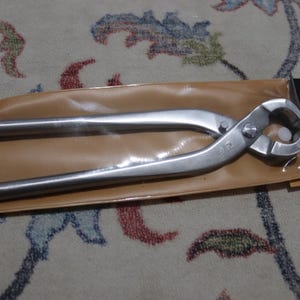 May include: A pair of stainless steel bonsai pliers, resting in a brown plastic sleeve. The pliers have long handles and a curved cutting head. A black box with Japanese text is visible.
