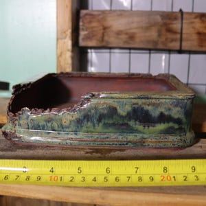 May include: A rectangular ceramic bonsai pot with a distressed edge. The pot has a dark green and blue glaze on the exterior and a reddish-brown interior. A yellow tape measure shows the pot is approximately 8 inches long.