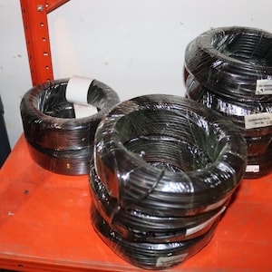 May include: Several coils of black wire are stacked on an orange shelf. The wire is wrapped in clear plastic. Some coils have white labels attached. A few coils of brown wire are visible on the right.