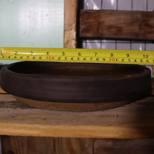 May include: A dark brown ceramic bowl, approximately 10 inches in diameter, is measured with a yellow tape measure. The bowl has a wide, rounded rim and sits on a wooden surface.