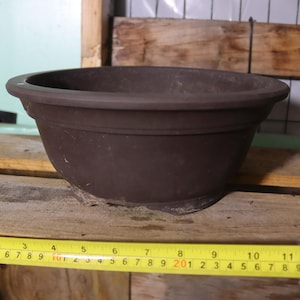 May include: A brown, round plant pot with a wide rim, approximately 11 inches in diameter. The pot sits on a wooden surface, with a measuring tape in the foreground. The pot is made of a dark brown material.