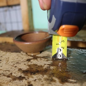 May include: A small, brown ceramic bowl is measured with a yellow tape measure. The bowl is approximately 2 inches deep. The tape measure is held by a person's hand. The bowl sits on a textured surface.