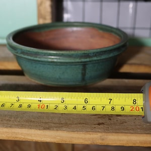 May include: A handmade, teal-colored ceramic bowl with a brown interior. The bowl is approximately 8 inches in diameter. A yellow tape measure is placed next to the bowl.