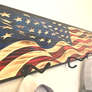 May include: Wooden wall-mounted coat rack featuring a carved American flag design. The flag has a blue field with white stars, red and white stripes, and a dark brown frame. Includes black metal hooks.
