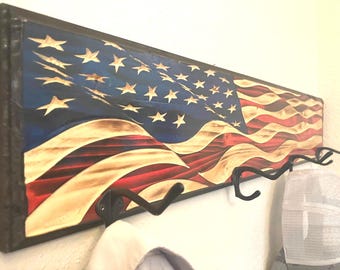 May include: Wooden wall-mounted coat rack featuring a carved American flag design. The flag has a blue field with white stars, red and white stripes, and a dark brown frame. Includes black metal hooks.