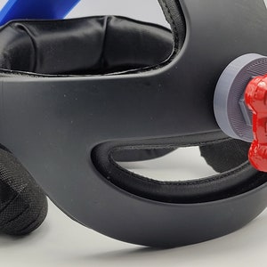 May include: A black VR headset with a red valve knob on the back. The headset has a black headband and a black foam padding around the eyes.