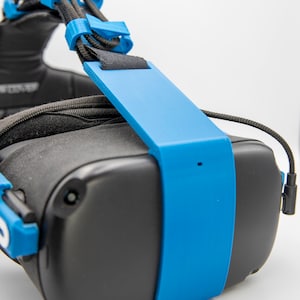 May include: A blue 3D printed head strap mount for a virtual reality headset. The strap is attached to the headset's faceplate and has a black strap that goes over the head.