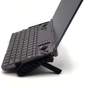 May include: A dark gray handheld gaming PC with an open screen. The device features a full keyboard, a touchpad, and gaming controls. It is propped up on a black stand, angled for viewing.
