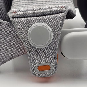 May include: Close-up of a gray and orange VR headset strap with a white button and a white oval-shaped cushion.