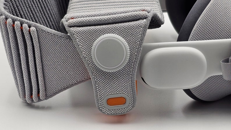 May include: Close-up of a gray and orange VR headset strap with a white button and a white oval-shaped cushion.
