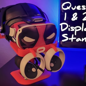 Op de afbeelding: Een rode en zwarte 3D-geprinte standaard voor Oculus Quest 1 en 2 VR-headsets. De standaard heeft twee slots voor controllers en een ruimte voor de headset. De standaard staat op een zwart oppervlak met een blauw licht dat er vanaf reflecteert.