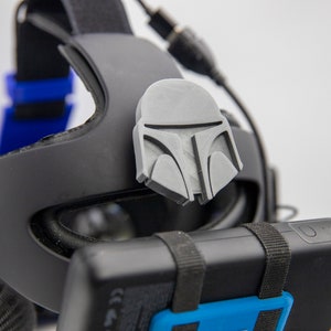 Könnte beinhalten: Ein schwarzes Virtual-Reality-Headset mit einem grauen, 3D-gedruckten Mandalorianer-Helm-Zubehör, das oben angebracht ist. Das Headset hat ein blaues Band und einen schwarzen Akku, der unten angebracht ist.