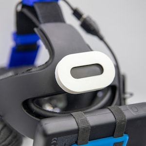 Może przedstawiać: Czarny zestaw słuchawkowy do wirtualnej rzeczywistości z białym czujnikiem z przodu. Zestaw słuchawkowy ma niebieski pasek i czarny akumulator przymocowany do boku.