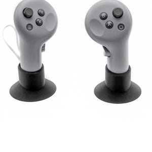 May include: Two gray gaming controllers with black buttons and a black base. Each controller has a rounded top with buttons labeled with letters and symbols. The controllers are designed for gaming.