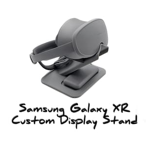 May include: A gray Samsung Galaxy XR custom display stand. The stand is designed to hold a VR headset. The stand has a base with a small platform and a curved support for the headset. The text "Samsung Galaxy XR Custom Display Stand" is written in black.