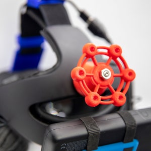 May include: A close-up of a red plastic knob with a silver screw in the center. The knob is attached to a black headband.