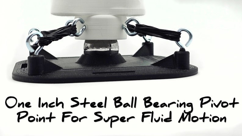 Puede incluir: Primer plano de una base negra con un punto de pivote de rodamiento de bolas de acero de 2,5 cm. La base tiene cuatro ganchos met&aacute;licos con cuerdas el&aacute;sticas negras unidas a ellos. "One Inch Steel Ball Bearing Pivot Point For Super Fluid Motion" est&aacute; escrito debajo de la base.