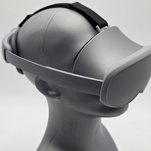May include: A gray VR headset with a black strap is displayed on a gray mannequin head. The headset has a curved design with a visor and adjustable side arms. The background is plain white.