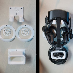 Puede incluir: Un soporte de pared blanco para auriculares con el logotipo de HP, diseñado para sostener un auricular de realidad virtual, controladores y cables. El soporte tiene un compartimento para los auriculares, dos compartimentos para los controladores y un compartimento para los cables.