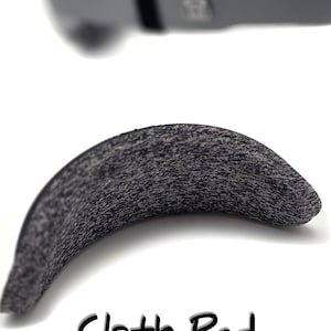May include: A curved, gray cloth pad with a textured surface. The text "Cloth Pad is Comfy & Washable" is written in black. The image also shows a blurred gray object in the background.