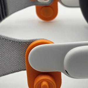 May include: Close-up of a gray headband with two orange plastic tabs. The tabs have a small, round button on the top. The text "Tab Connection Style" is written below the headband.