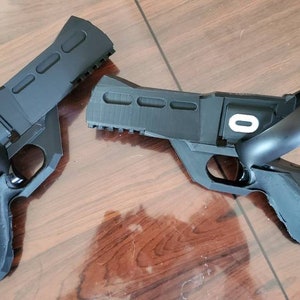 Könnte beinhalten: Zwei schwarze VR-Pistolen-Controller mit weißem "O"-Logo, die das Virtual-Reality-Spielerlebnis verbessern sollen. Die Controller liegen auf einer Holzoberfläche.