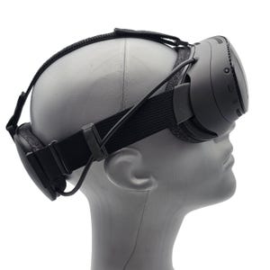 May include: A black virtual reality headset with an adjustable elastic strap system and a padded rear head support, positioned on a gray mannequin head. The device features a sleek front casing with visible side buttons and a connecting cable.