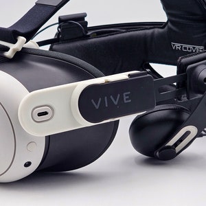 Puede incluir: Un auricular de realidad virtual blanco y negro con una correa negra y la palabra "VIVE" impresa en el lateral. El auricular tiene una correa de audio negra con un auricular negro en el lado derecho.