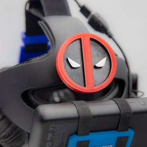 Peut inclure: Un casque de réalité virtuelle noir avec une plaque frontale Deadpool rouge et noire.