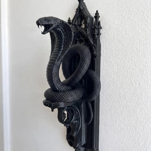 以下が含まれることがあります： マットブラックの壁掛け棚で、フードを広げて口を開けたコブラが巻き付いています。棚は、複雑な尖頭アーチと装飾的な尖塔が施された垂直のバックプレートによって支えられており、建築的なデザインが特徴です。