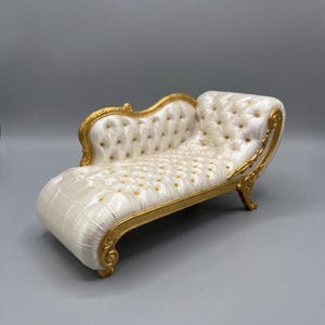 May include: A miniature chaise lounge featuring a pearlescent white tufted seat with gold button accents. The frame is constructed from ornate gold-painted material with carved scrollwork details along the backrest, arm, and base legs.