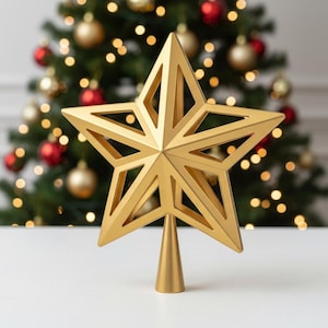 May include: A gold star tree topper with a geometric design. The star is set against a blurred background of a Christmas tree decorated with red and gold ornaments and lights. The star is a festive holiday decoration.