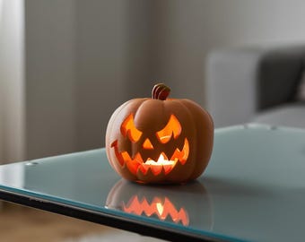 Może przedstawiać: Matowa, pomarańczowa dynia z wydruku 3D z wyciętym, poszarpanym, złowrogim wyrazem twarzy. Puste wnętrze zawiera źródło światła, które oświetla wycięcia, rzucając ciepłe światło przez oczy i usta.